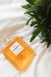 A glass bottle of Chanel No. 5 eau de parfum rests on a white fabric surface next to green foliage. The bottle contains an amber-colored liquid and features a rectangular shape with a minimalist label.