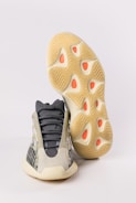 A pair of athletic shoes with a futuristic design. The upper part of the shoe features a mix of dark gray and beige with mesh panels, giving it a sleek appearance. The sole is thick and has a unique wavy pattern with orange accents.