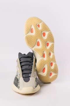 A pair of athletic shoes with a futuristic design. The upper part of the shoe features a mix of dark gray and beige with mesh panels, giving it a sleek appearance. The sole is thick and has a unique wavy pattern with orange accents.
