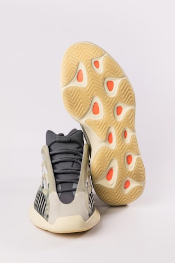 A pair of athletic shoes with a futuristic design. The upper part of the shoe features a mix of dark gray and beige with mesh panels, giving it a sleek appearance. The sole is thick and has a unique wavy pattern with orange accents.