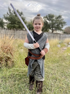A young girl stands outdoors holding a toy lightsaber. She is dressed in a costume reminiscent of a Jedi, with a gray scarf-like garment, brown belt, and brown boots. The background consists of tall grass, a wooden fence, and leafy trees under a cloudy sky.