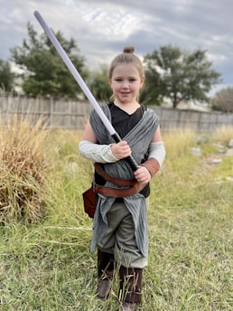 A young girl stands outdoors holding a toy lightsaber. She is dressed in a costume reminiscent of a Jedi, with a gray scarf-like garment, brown belt, and brown boots. The background consists of tall grass, a wooden fence, and leafy trees under a cloudy sky.