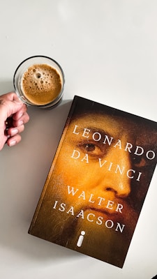 A person holding a cup of coffee is positioned next to a book titled 'Leonardo Da Vinci' by Walter Isaacson. The cover features a close-up image of a face with a golden hue. The background is a plain, light-colored surface.