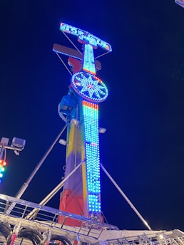 A brightly lit amusement park ride towering against the night sky, adorned with vivid blue and red lights forming patterns and shapes. The structure is metallic, with supporting beams and a safety railing visible near the base.