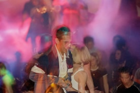 A lively scene in a dimly lit club with a group of young people. The atmosphere is filled with vibrant colors and hazy smoke, suggesting a party or dance event. A man and a woman are in the foreground, with the man smiling and the woman standing close to him. The background reveals several other individuals mingling and enjoying the environment.