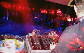 A DJ operates a mixing console in a dimly lit setting, surrounded by vibrant red and blue lights. The turntable features artwork of a woman's face. In the background, a crowd is visible, creating an energetic and lively atmosphere.