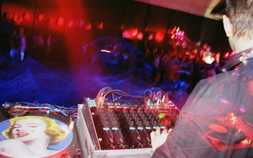 A DJ operates a mixing console in a dimly lit setting, surrounded by vibrant red and blue lights. The turntable features artwork of a woman's face. In the background, a crowd is visible, creating an energetic and lively atmosphere.