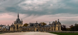 A grand, historic chateau with intricate architectural details is situated in a serene landscape. The expansive building showcases multiple spires and ornate towers. The scene is complemented by a cloudy sky that casts a dramatic atmosphere over the chateau, and surrounding trees display autumnal colors.