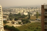 A densely built residential area is visible with multiple apartment buildings arranged in rows. The foreground features greenery and undeveloped land, while a clear road separates the open area from the buildings. Haze in the background suggests pollution or mist.