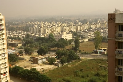 A densely built residential area is visible with multiple apartment buildings arranged in rows. The foreground features greenery and undeveloped land, while a clear road separates the open area from the buildings. Haze in the background suggests pollution or mist.