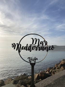 A decorative metal sign with the words 'Mar Mediterráneo Tarifa' is positioned in front of a scenic seascape. The sky is partly cloudy, and the sea is calm, with rocks lining the shore.