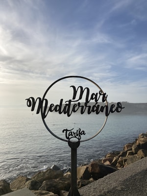 A decorative metal sign with the words 'Mar Mediterráneo Tarifa' is positioned in front of a scenic seascape. The sky is partly cloudy, and the sea is calm, with rocks lining the shore.