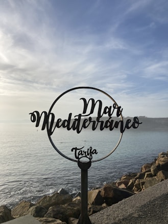 A decorative metal sign with the words 'Mar Mediterr&aacute;neo Tarifa' is positioned in front of a scenic seascape. The sky is partly cloudy, and the sea is calm, with rocks lining the shore.
