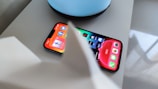 A smartphone displaying various app icons on its screen, placed on a light-colored surface near a blue circular object. Part of a white object is partially visible in the foreground.