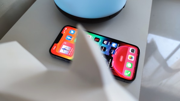 A smartphone displaying various app icons on its screen, placed on a light-colored surface near a blue circular object. Part of a white object is partially visible in the foreground.