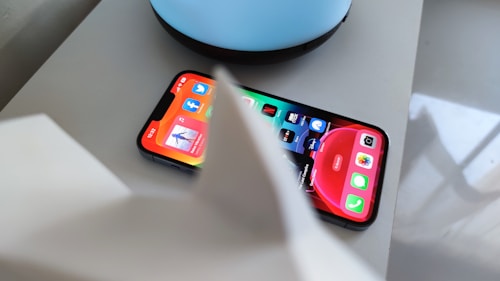 A smartphone displaying various app icons on its screen, placed on a light-colored surface near a blue circular object. Part of a white object is partially visible in the foreground.