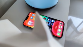 A smartphone displaying various app icons on its screen, placed on a light-colored surface near a blue circular object. Part of a white object is partially visible in the foreground.