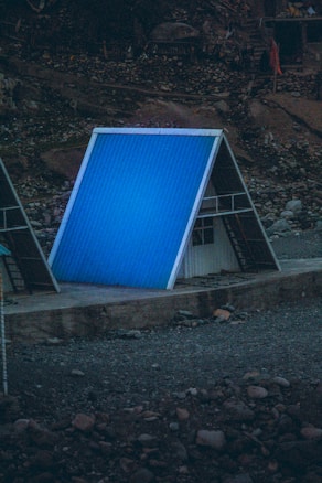 An A-frame structure with a bright blue metal roof is situated on a rocky, barren landscape. The building appears to have a small entrance area beneath the roof, possibly used for storage or temporary shelter.