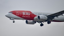 A commercial airplane in flight with a red and white paint scheme, featuring branding for RIU Hotels & Resorts, and websites TUI.COM. The airplane is captured in mid-air with its landing gear extended against a cloudy sky.
