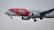 A commercial airplane in flight with a red and white paint scheme, featuring branding for RIU Hotels & Resorts, and websites TUI.COM. The airplane is captured in mid-air with its landing gear extended against a cloudy sky.
