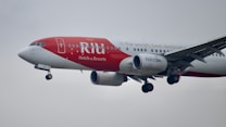 A commercial airplane in flight with a red and white paint scheme, featuring branding for RIU Hotels & Resorts, and websites TUI.COM. The airplane is captured in mid-air with its landing gear extended against a cloudy sky.