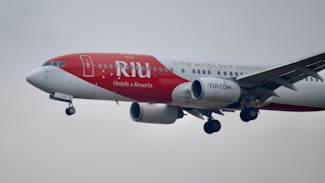 A commercial airplane in flight with a red and white paint scheme, featuring branding for RIU Hotels & Resorts, and websites TUI.COM. The airplane is captured in mid-air with its landing gear extended against a cloudy sky.