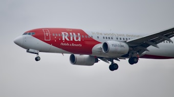 A commercial airplane in flight with a red and white paint scheme, featuring branding for RIU Hotels & Resorts, and websites TUI.COM. The airplane is captured in mid-air with its landing gear extended against a cloudy sky.