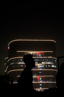 A modern, multi-story building is illuminated with soft yellow lights against a dark night sky. The lights outline the building's curved architecture. In the foreground, there is a silhouette of a person or object, adding a sense of depth and contrast to the scene.