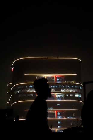A modern, multi-story building is illuminated with soft yellow lights against a dark night sky. The lights outline the building's curved architecture. In the foreground, there is a silhouette of a person or object, adding a sense of depth and contrast to the scene.