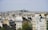 A panoramic view of a dense urban cityscape featuring numerous buildings with white and beige facades and traditional Parisian rooftops. In the background, atop a hill, a prominent basilica with its notable dome is surrounded by greenery.