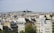 A panoramic view of a dense urban cityscape featuring numerous buildings with white and beige facades and traditional Parisian rooftops. In the background, atop a hill, a prominent basilica with its notable dome is surrounded by greenery.
