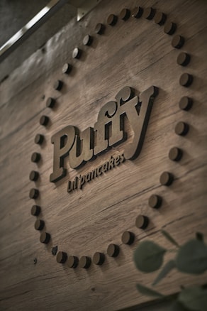 A wooden background features a circular pattern of black dots surrounding the raised, bold lettering that reads 'Puffy lil' pancakes'. The typography is stylish and the setting is industrial with a natural, rustic feel. The image is angled, focusing on the brand name prominently.