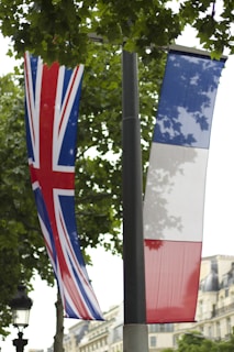 Two flags, one of the United Kingdom with the Union Jack design, and the other of France with the blue, white, and red tricolor, hang from a pole. They are set against a backdrop of leafy trees and some European-style buildings in the background.