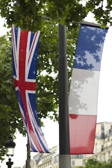 Two flags, one of the United Kingdom with the Union Jack design, and the other of France with the blue, white, and red tricolor, hang from a pole. They are set against a backdrop of leafy trees and some European-style buildings in the background.