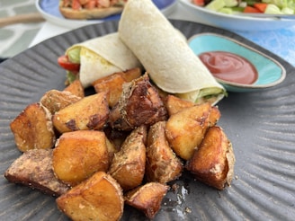 A close-up of a plated meal featuring golden-brown roasted potato wedges and a tortilla wrap filled with vegetables. The plate also includes a small dish of red sauce. The background shows blurred images of other food items on different plates.