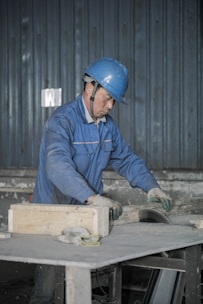 A person wearing a blue hard hat and a blue work uniform is engaged in work, handling a piece of equipment or material on a table in an industrial setting. The background features a corrugated metal wall.