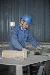 A person wearing a blue hard hat and a blue work uniform is engaged in work, handling a piece of equipment or material on a table in an industrial setting. The background features a corrugated metal wall.