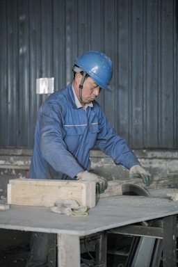 A person wearing a blue hard hat and a blue work uniform is engaged in work, handling a piece of equipment or material on a table in an industrial setting. The background features a corrugated metal wall.