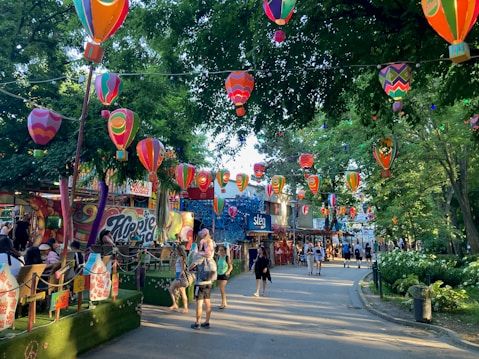 A vibrant outdoor scene with numerous colorful hot air balloon decorations hanging from trees creating a festive atmosphere. People walk along a pathway lined with greenery, colorful graffiti, and stalls or booths. Brightly colored signs and decorations enhance the lively ambiance of the area.