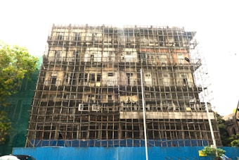 A multi-story building is undergoing construction or renovation, with a complex network of scaffolding covering its facade. The structure appears old and possibly in need of repair. A blue barrier is set up at the base, indicating a construction zone. A tree is visible on the left side of the image.