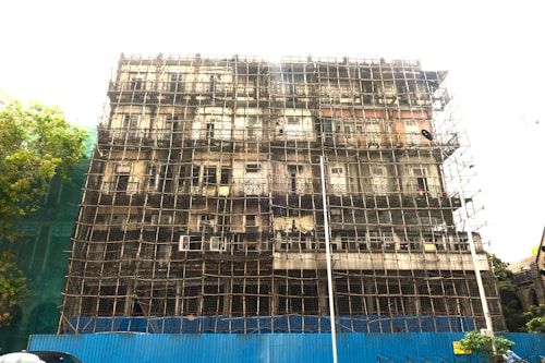 A multi-story building is undergoing construction or renovation, with a complex network of scaffolding covering its facade. The structure appears old and possibly in need of repair. A blue barrier is set up at the base, indicating a construction zone. A tree is visible on the left side of the image.