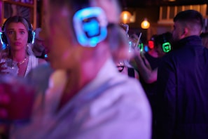 A lively atmosphere with multiple people wearing illuminated headphones, suggesting a silent disco or party setting. One person in the foreground holds a drink, while others are animatedly socializing in the background, all under dim, colorful lighting.