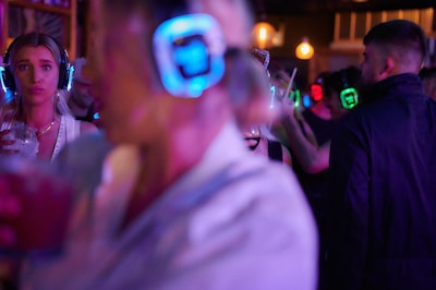 A lively atmosphere with multiple people wearing illuminated headphones, suggesting a silent disco or party setting. One person in the foreground holds a drink, while others are animatedly socializing in the background, all under dim, colorful lighting.