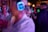A lively atmosphere with multiple people wearing illuminated headphones, suggesting a silent disco or party setting. One person in the foreground holds a drink, while others are animatedly socializing in the background, all under dim, colorful lighting.