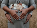 A close-up of a pregnant belly with two hands gently cradling it, forming a heart shape. The person is wearing a dress with a floral belt featuring beige, white, and brown flowers.