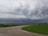 Dark storm clouds loom over a rural landscape with green fields and dirt roads. The sky exhibits a dramatic and potentially ominous atmosphere, with power lines running across the scene.