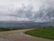 Dark storm clouds loom over a rural landscape with green fields and dirt roads. The sky exhibits a dramatic and potentially ominous atmosphere, with power lines running across the scene.