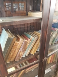 An antique wooden bookcase with glass doors holds a collection of old, worn books with varying cover colors and visible signs of aging. Some books have faded or torn spines, and dust is present on the shelves, indicating infrequent use.