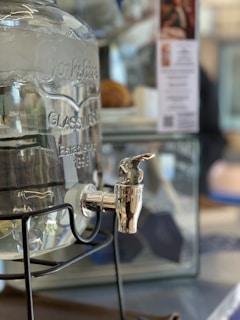 A large glass beverage dispenser with an engraved design, including text that reads 'Established 1840.' The dispenser is mounted on a black metal stand, with a shiny silver spigot at the bottom right. In the background, items such as a blurred menu and a geometric patterned object are visible.