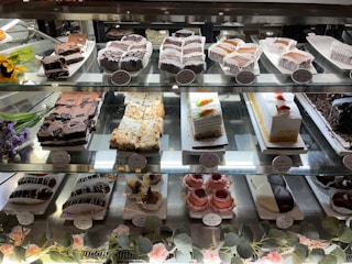 A display case filled with various desserts, including squares of brownies, slices of cakes, and pastries, all neatly arranged on glass shelves. There are labels in front of each item indicating the price. Flowers and green leaves are used as decorative elements in the bottom section of the case.